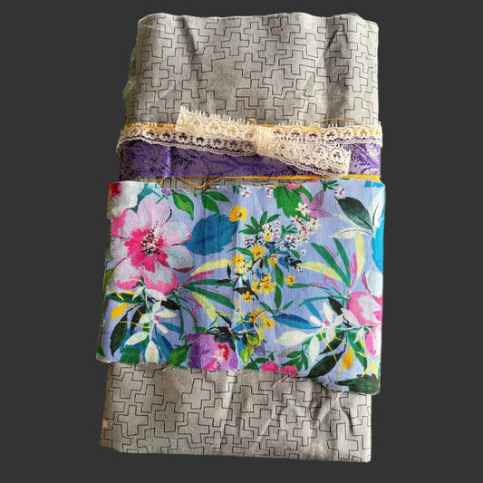 A folded fabric bundle or gift wrap featuring a gray geometric patterned fabric, a purple lace trim, a cream lace ribbon, and a central tropical floral print panel with pink, blue, yellow, and green.